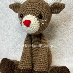 May include: A handmade crochet reindeer with brown and cream yarn. The reindeer has a red nose, black eyes, and white heart-shaped antlers. The reindeer is sitting with its legs extended. The text "&copy;ByHerHandBoutique" is visible.