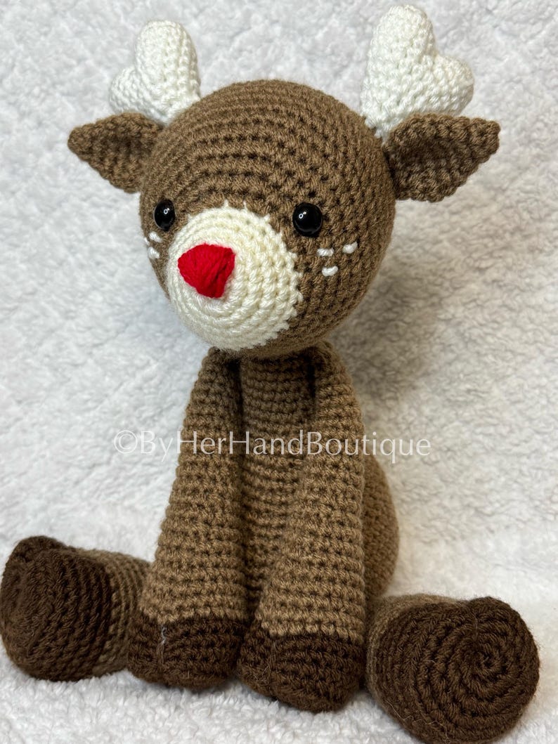 May include: A handmade crochet reindeer with brown and cream yarn. The reindeer has a red nose, black eyes, and white heart-shaped antlers. The reindeer is sitting with its legs extended. The text "&copy;ByHerHandBoutique" is visible.
