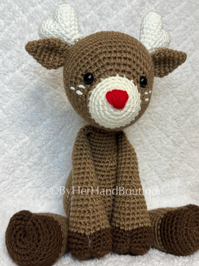 May include: A handmade crochet reindeer with brown and white accents. The reindeer has a brown body, legs, and head, with white antlers and a white muzzle. It features a red nose and black eyes. The text "&copy;ByHerHandBoutique" is visible.