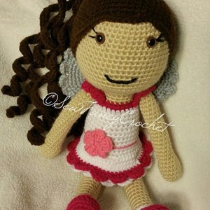 May include: A handmade crochet doll with brown curly hair, a pink flower, and a white and red dress. The doll has gray wings and pink shoes. The doll is made of yarn and is a whimsical toy.