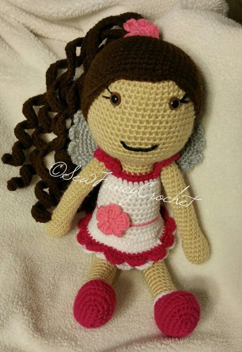 May include: A handmade crochet doll with brown curly hair, a pink flower, and a white and red dress. The doll has gray wings and pink shoes. The doll is made of yarn and is a whimsical toy.