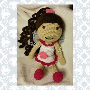 May include: A handmade crochet doll with brown curly hair, a pink flower, and small gray wings. The doll wears a white dress with pink trim and a pink flower. The doll's feet are pink. The text "Sew Fancy Crochet" is visible.