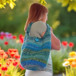 Peut inclure: Un sac à bandoulière crocheté bleu et vert avec une fermeture à gros bouton bleu. Le sac présente des rayures horizontales de différentes nuances de bleu et de vert. Le sac est porté à l'extérieur dans un jardin.