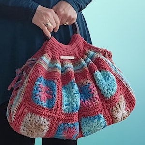 May include: A handmade, coral-colored crochet bag with a wooden handle. The bag features square panels with blue, pink, and tan floral designs. The bag is held by a person wearing a blue dress.