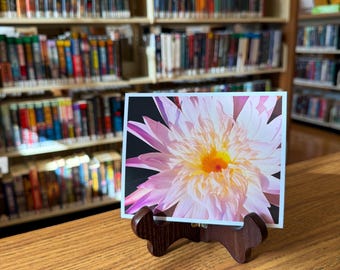 Può includere: Una stampa rettangolare raffigurante un vivace fiore di loto rosa e bianco con un centro giallo brillante, esposta su un cavalletto in legno marrone scuro. I petali del fiore sono stratificati e dettagliati, dal centro.