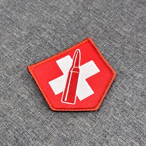 Puede incluir: Un parche bordado pentagonal con un fondo rojo brillante y un símbolo de cruz blanca en el centro. Un gráfico de bala con contorno blanco está colocado verticalmente sobre la cruz, creando un diseño de alto contraste con bordes cosidos.