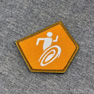 Puede incluir: Un parche bordado pentagonal con un fondo naranja vibrante y un borde cosido de color marrón oscuro. Centrado en el parche hay un icono gráfico blanco que representa una figura estilizada en movimiento, corriendo o girando.