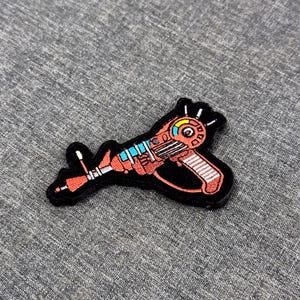 Puede incluir: Un parche bordado que muestra una pistola de rayos de estilo retro en tonos naranja, rojo y azul claro. El diseño incluye un indicador circular con un segmento amarillo, un mango a rayas y una boquilla puntiaguda, todo con un borde negro.