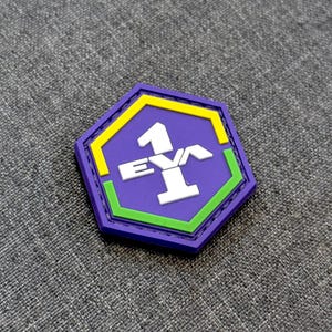 Puede incluir: Un parche hexagonal con fondo morado y borde amarillo y verde. En el centro aparece el texto blanco EVA 1 en una fuente gruesa y geométrica. El diseño está en relieve y fabricado con un material liso y resistente.