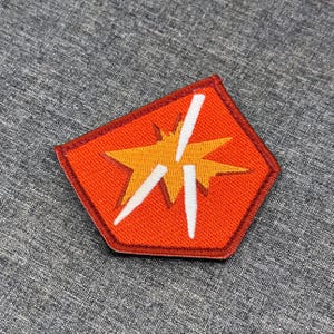 Puede incluir: Un parche bordado pentagonal con un fondo naranja vibrante y un borde cosido en rojo oscuro. El centro muestra un gráfico de estrella amarilla con tres formas blancas puntiagudas similares a rayos superpuestas.