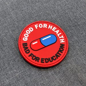 Puede incluir: Un parche circular rojo con un gráfico central de una pastilla dividida en mitades roja y azul con un reflejo blanco. El texto GOOD FOR HEALTH está impreso en blanco en la parte superior y BAD FOR EDUCATION en negro en la parte inferior.
