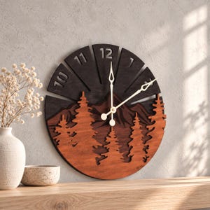 Könnte beinhalten: Eine runde Wanduhr aus Holz mit einer geschichteten Berg- und Tannenlandschaft in Braun- und Schwarztönen. Das Zifferblatt zeigt die Zahlen 10, 11, 12 und 1, mit dünnen, hellen Metallzeigern, die die Uhrzeit anzeigen.