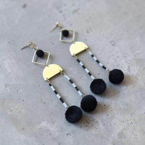 Può includere: Orecchini pendenti con una forma a rombo color oro, un semicerchio color oro e due fili di perline bianche e nere. Ogni filo termina con un piccolo pon-pon nero, e un pon-pon nero è posizionato al centro della cornice a rombo.