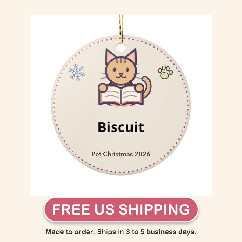 May include: A round, cream-colored ceramic ornament featuring a brown tabby cat reading an open book. The design includes a blue snowflake, a paw print, and the text "Biscuit" and "Pet Christmas 2026" in black, with a gold hanging cord.