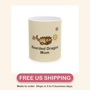 May include: A beige ceramic mug featuring a cartoon illustration of a brown bearded dragon, a small orange leaf, a yellow sun, and a pink flower. The text "Bearded Dragon Mom" is printed in dark brown letters below the lizard graphic.