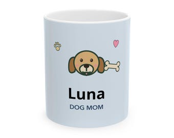 May include: A light blue ceramic mug featuring a cartoon dog face, a bone, a paw print, and a small pink heart. Below the dog graphic, the text reads Luna in large black letters and DOG MOM in smaller dark blue capital letters.