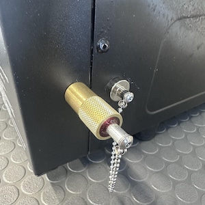 May include: A black metal housing features a cylindrical brass-colored component with a textured knurled grip. A silver-toned metal pin is inserted into the center, attached to a small silver ball chain that hangs downward from the device.