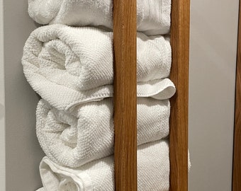 May include: A wooden towel rack with four white towels stacked on top of each other and three more towels rolled up in a small shelf below. The rack is made of light brown wood with a natural finish.
