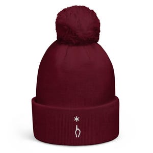Könnte beinhalten: Eine tiefburgunderrote Strickmütze mit passendem Bommel und umgeschlagenem Saum. Auf dem Saum ist ein kleines weißes Sternchen eingestickt, das sich direkt über einer weißen Grafik einer nach oben zeigenden Hand befindet.