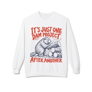 Könnte beinhalten: Ein weißes Langarm-Sweatshirt mit einer Grafik eines Bibers, der an einem Holzstapel arbeitet. Der rote Text über und unter der Illustration lautet, IT'S JUST ONE DAM PROJECT AFTER ANOTHER, in einer fetten, serifenlosen Schrift.
