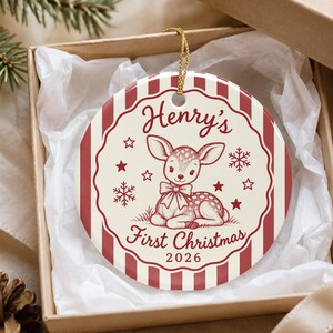 May include: A round ceramic Christmas ornament featuring a red and white striped border. The center displays a fawn illustration with a bow, surrounded by stars and snowflakes. Text reads: Henry's First Christmas 2026.