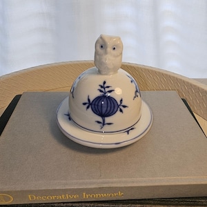 May include: A white porcelain butter dish with a dome-shaped lid topped by a sculpted owl figurine. The lid and base are decorated with a blue onion pattern featuring stylized floral motifs and thin blue lines along the edges.