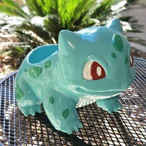 May include: A light blue ceramic planter shaped like the character Bulbasaur, featuring dark green spots on its back and sides, white and red eyes, and a hollow opening on its back for holding small plants or decorative items.