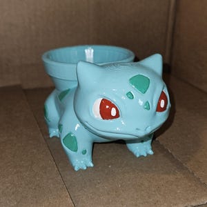 May include: A light blue planter shaped like the character Bulbasaur, featuring dark green spots on its body, red eyes, and a wide circular opening on its back for holding small plants or decorative items.