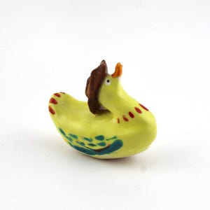 May include: A small yellow ceramic duck figurine featuring a brown hat on its head, an orange beak, and painted details including red markings on the wings and back, along with blue and green patterns on the sides of its body.