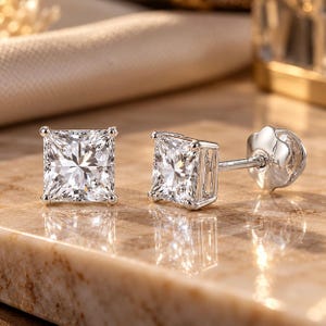 May include: A pair of silver-toned stud earrings featuring square-cut clear stones set in four-prong baskets. One earring is displayed from the front, while the other is shown from the side, revealing the post and a scalloped-edge backing.