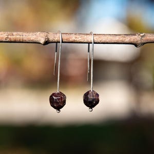 May include: A pair of drop earrings featuring faceted, dark red, spherical beads suspended from long, thin, silver-toned metal hooks. Each bead is secured at the base by a small, round silver-toned metal accent, creating a simple, linear design.