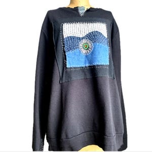 May include: A black long-sleeve sweatshirt featuring a square patch on the chest with white stitched lines, a blue fabric section, and a central flower graphic with white petals and a green center. The neckline has a small frayed fabric detail.