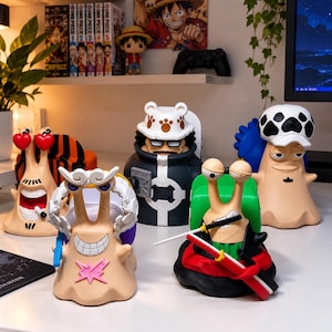 Può includere: Una collezione di cinque statuette a forma di lumaca di One Piece. Le figure presentano vari design di personaggi, tra cui uno con occhi a forma di cuore, uno con un cappello con orecchie da orso, uno con una spada e uno con un cappello a pois, su una superficie bianca.