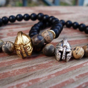 May include: Two beaded bracelets with a black onyx and brown jasper stone design. Each bracelet features a metal helmet charm, one gold and one silver.
