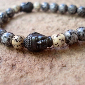 May include: A black beaded bracelet with a silver Buddha head charm. The beads are speckled with white and gray.