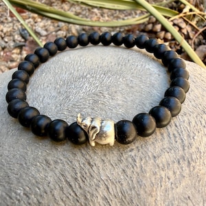 May include: A stretch bracelet composed of matte black spherical beads arranged in a circular pattern. The bracelet features a single silver-toned metal charm shaped like an elephant, positioned between two of the black beads as a focal point.