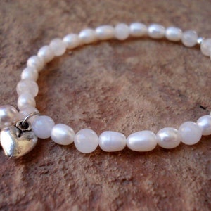 May include: A white pearl bracelet with a silver heart charm and angel wings. The bracelet is made with oval-shaped pearls and a silver clasp.