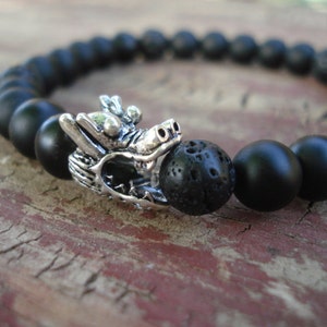 May include: Black beaded bracelet with a silver dragon charm and a black lava rock bead.