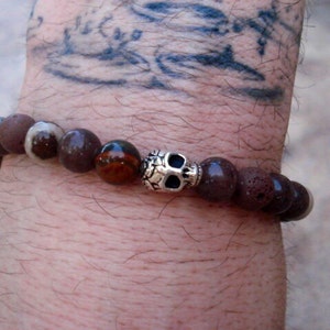 Puede incluir: Un brazalete de cuentas marrones y blancas con un dije de calavera de plata. El brazalete está hecho de piedras naturales y cuentas de lava.
