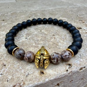 May include: A black beaded bracelet with a gold-colored Spartan helmet charm. The bracelet features matte black beads, speckled brown beads, and gold-colored spacers. The bracelet is displayed on a textured surface.