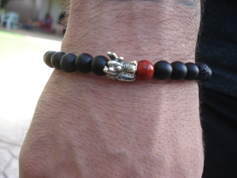 May include: Black beaded bracelet with a silver dragon charm and a red bead. The bracelet is made of lava rock and onyx beads.