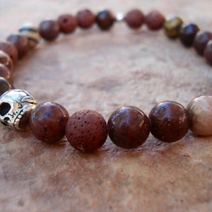 Puede incluir: Un brazalete de cuentas marrones y beige con un dije de calavera de plata. El brazalete está hecho con roca volcánica, ojo de tigre y otras piedras naturales.