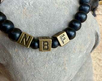 May include: A stretch bracelet featuring matte black spherical beads and three square metallic bronze-toned beads. The square beads are embossed with the letters N, B, and F in a dark, recessed font, arranged centrally along the circular bead strand.