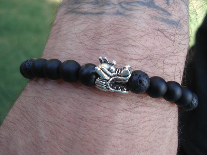 May include: Black beaded bracelet with a silver dragon charm. The beads are made of lava rock and onyx.