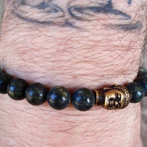 May include: A dark green beaded bracelet with a gold Buddha head charm. The bracelet is on a person's wrist.