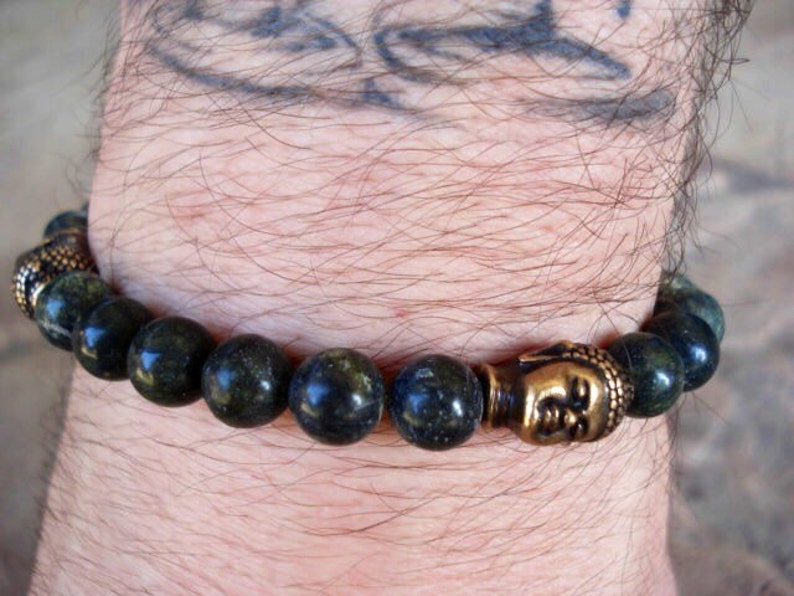 May include: A dark green beaded bracelet with a gold Buddha head charm. The bracelet is on a person's wrist.