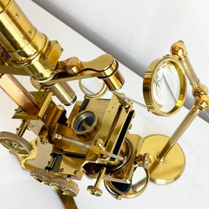 May include: A brass microscope featuring a central stage, multiple adjustment knobs, and a vertical eyepiece tube. An attached magnifying lens on a jointed arm extends from the side, positioned near the circular stage and reflective mirror.