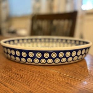 May include: A round ceramic baking dish featuring a white interior and exterior decorated with a repeating pattern of blue and white concentric circles with small brown centers. The dish has a low profile and a smooth, glazed finish.