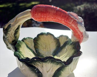 May include: A ceramic basket shaped like a head of cabbage with green and cream leaves. The handle is formed by a large orange carrot with a white rabbit head attached to one side, featuring painted details and a glossy finish.
