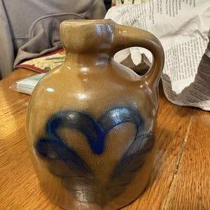 May include: A tan stoneware jug with a single handle and a narrow neck. The body of the jug features a hand-painted cobalt blue floral design centered on the front. The surface has a smooth, glossy glaze finish over the tan clay body.